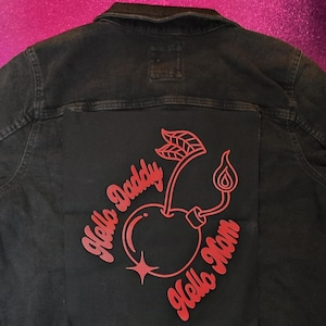May include: A black denim jacket featuring a large red graphic on the back. The design depicts a cherry with a lit fuse, accompanied by the text Hello Daddy and Hello Mom in a stylized script font, centered on the back panel of the garment.