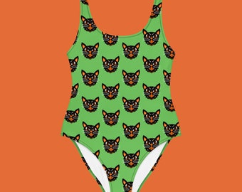 Op de afbeelding: Een badpak met een herhaald patroon van zwarte kattengezichten op een felgroene achtergrond. De katten hebben oranje ogen en een strenge uitdrukking. Het badpak heeft een ronde hals en een hoog uitgesneden been.