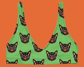 Op de afbeelding: Een groene bikinitop met een herhaald patroon van zwarte kattengezichten met oranje accenten. De kattengezichten hebben witte snorharen en een boze uitdrukking. De top heeft een V-hals en staat tegen een oranje achtergrond.