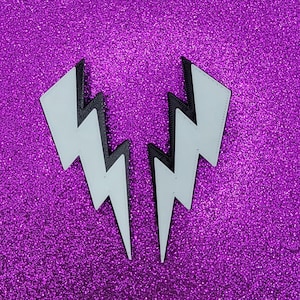 Könnte beinhalten: Zwei weiße, blitzförmige Accessoires mit schwarzem Rand, vor einem glitzernden lila Hintergrund. Die Blitze sind symmetrisch und scheinen aus einem glatten Material zu bestehen. Das Bild ist gut beleuchtet und hebt den Kontrast zwischen Weiß und Schwarz hervor.
