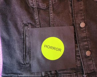May include: Black denim jacket with silver buttons and a small pocket. A black square with a neon green circle that says "HORROR" is attached to the jacket.