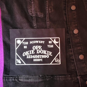 May include: Black denim jacket with a white Ouija board patch. The patch features the words "Yeah Midwest No Yeah No Ope Okie Dokie" and numbers 1-0. The jacket has metal buttons and a purple background.