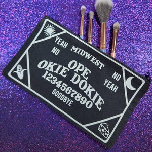 May include: Black makeup bag with a white "Ouija board" design featuring the text "Ope Dokie" and numbers 1234567890. The bag has a white sun and moon design on either side. The bag is lying on a purple sparkly surface with four makeup brushes sticking out of the top.