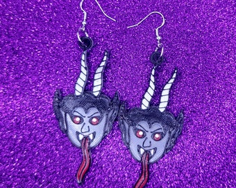 May include: A pair of dangle earrings featuring a cartoon devil head with red eyes, a black face, and a red tongue. The earrings are silver and have a black hook.