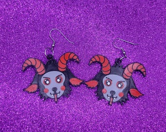 May include: A pair of dangle earrings featuring a cartoon devil goat with red horns, black fur, and a pink tongue sticking out. The earrings are made of black and red plastic and have silver hooks.