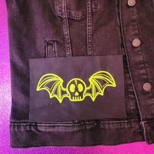 May include: A black denim jacket with a neon green bat-skull patch on the pocket. The patch features a skull with bat wings. The jacket has silver buttons and a classic design.