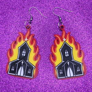 May include: A pair of black and yellow church earrings with flames. The church is black with white windows and a white cross on the roof. The flames are yellow and red.