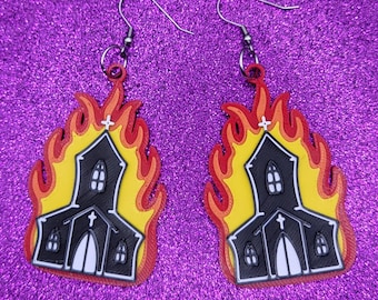 May include: A pair of black and yellow church earrings with flames. The church is black with white windows and a white cross on the roof. The flames are yellow and red.