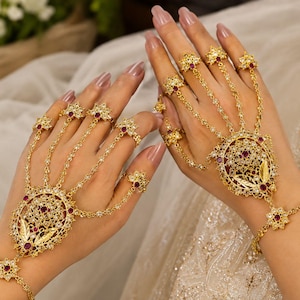 Puede incluir: Un par de joyas de mano doradas con intrincados diseños de filigrana, incrustadas con pequeños cristales transparentes y piedras de color rojo intenso. Cada pieza consta de un medallón central conectado por cadenas a varios anillos.