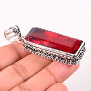 May include: A rectangular pendant featuring a deep red faceted gemstone set in a silver frame. The silver setting displays intricate filigree scrollwork along the sides and includes a polished silver bail at the top for attachment to a chain.