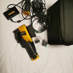 May include: A yellow and black handheld thermal imaging camera with a black carrying case, a black power adapter, and a black battery pack. The camera has a black handle and a yellow trigger. The camera is labeled "FLUKE" and "TI300".