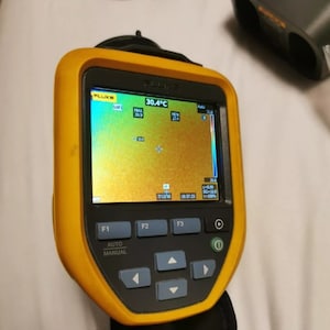 May include: A yellow and black handheld thermal imaging camera with a digital display showing a temperature reading of 30.4 degrees Celsius. The display shows a thermal image of a surface with a color scale ranging from blue to red. The camera has buttons labeled F1, F2, F3, Auto, and Manual.