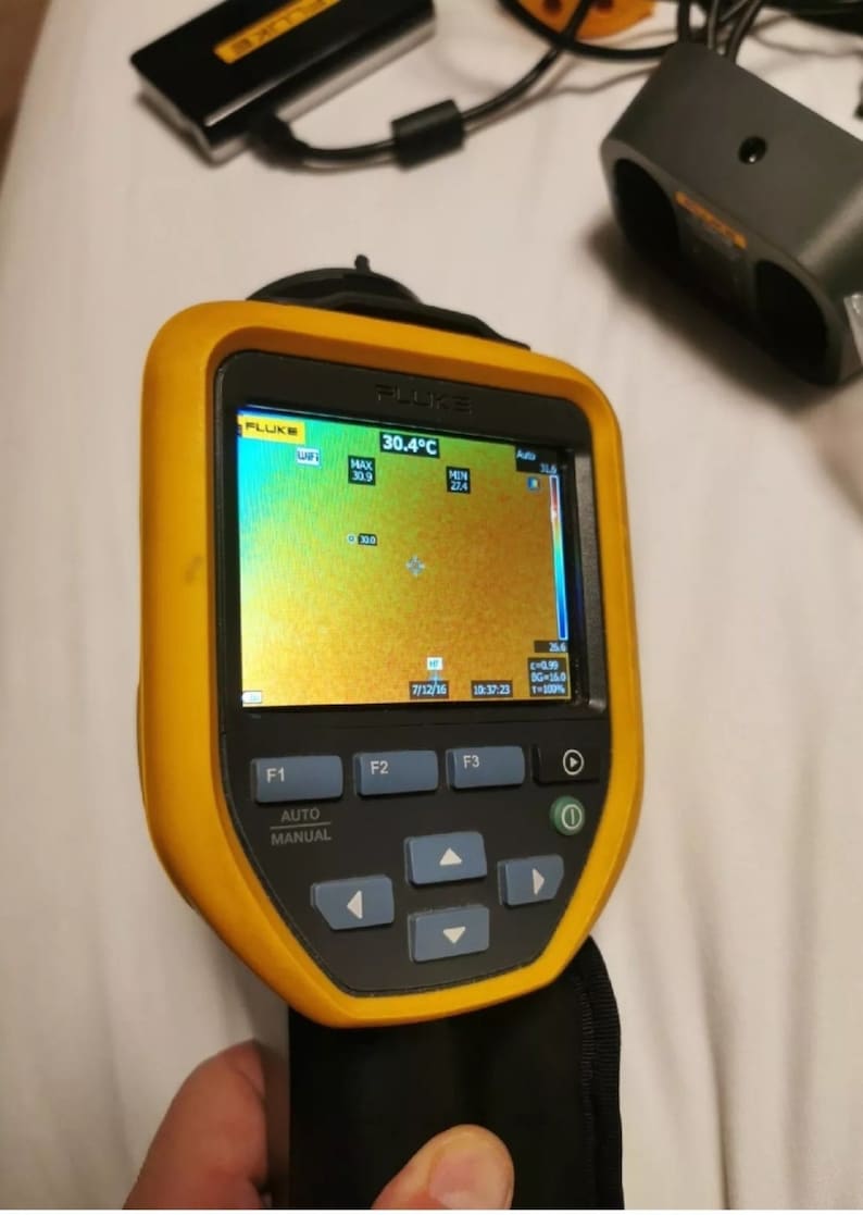 May include: A yellow and black handheld thermal imaging camera with a digital display showing a temperature reading of 30.4 degrees Celsius. The display shows a thermal image of a surface with a color scale ranging from blue to red. The camera has buttons labeled F1, F2, F3, Auto, and Manual.