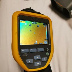 May include: A yellow and black handheld thermal imaging camera with a digital display showing a temperature reading of 30.4 degrees Celsius. The display shows a thermal image with a color scale ranging from blue to red. The camera has buttons labeled F1, F2, F3, Auto, and Manual.
