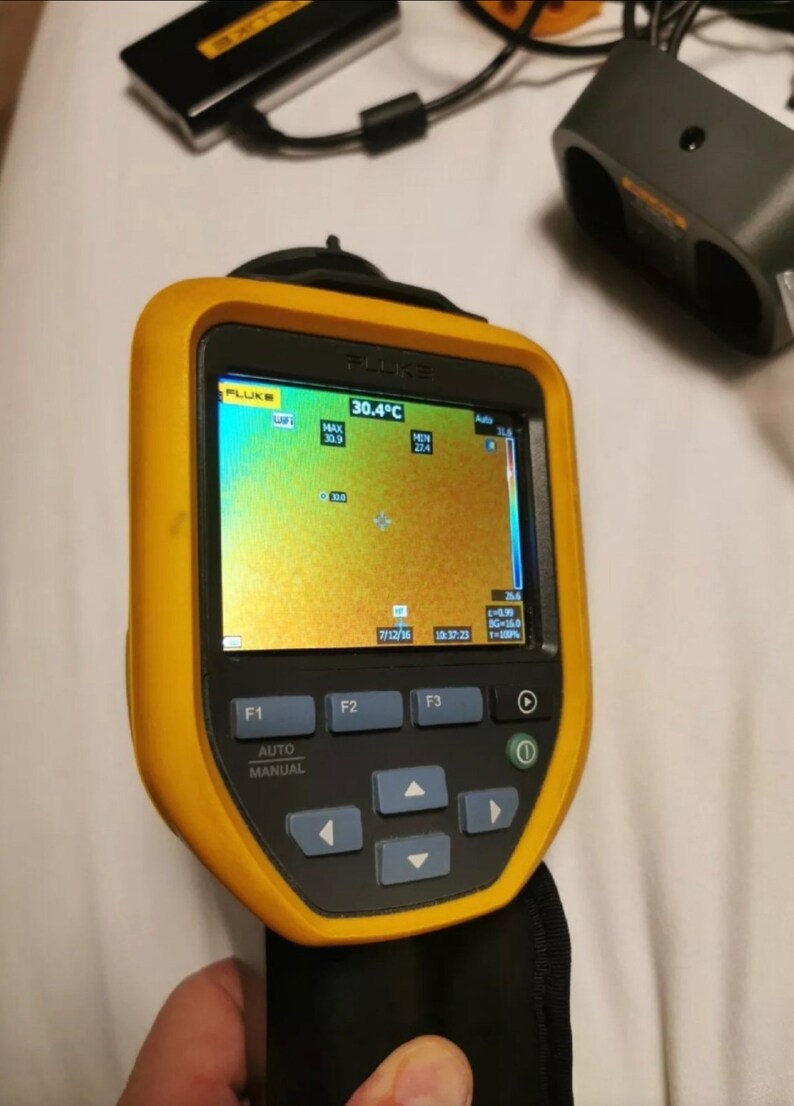 May include: A yellow and black handheld thermal imaging camera with a digital display showing a temperature reading of 30.4 degrees Celsius. The display shows a thermal image with a color scale ranging from blue to red. The camera has buttons labeled F1, F2, F3, Auto, and Manual.