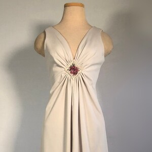 Pode incluir: Um vestido longo maxi, sem mangas, de cor cinza claro, com decote em V e uma pequena flor rosa como adorno no decote.