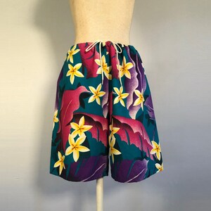 May include: Mid-length shorts featuring a vibrant tropical print with yellow plumeria flowers and large purple and magenta leaves against a deep teal background. The shorts have an elasticized waistband with a white drawstring cord at the center.