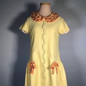 May include: A yellow vintage dress with a floral print collar and matching bows on the skirt. The dress has a button-up front and short sleeves.