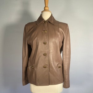 May include: A taupe leather jacket with a classic collar and four decorative gold buttons. The jacket features two front pockets with matching gold button details. The jacket is displayed on a mannequin.
