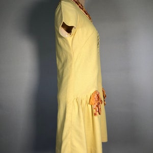 May include: A yellow dress with a short sleeve and a ruffled collar. The dress has a bow detail on the skirt.