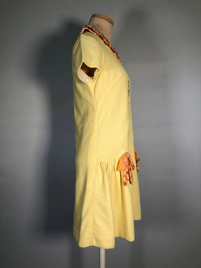 May include: A yellow dress with a short sleeve and a ruffled collar. The dress has a bow detail on the skirt.