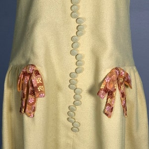 May include: A pale yellow dress with a row of white buttons down the front. The dress has two pockets with pink and brown embroidered bows on each side.