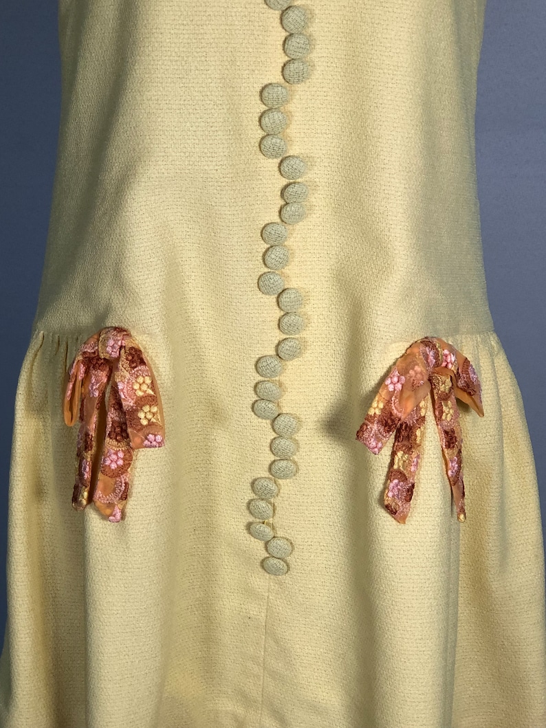 May include: A pale yellow dress with a row of white buttons down the front. The dress has two pockets with pink and brown embroidered bows on each side.