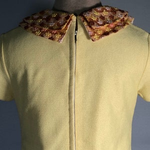 May include: A yellow short-sleeved dress with a large, embroidered collar. The collar is a multi-colored floral pattern in shades of pink, orange, and brown.