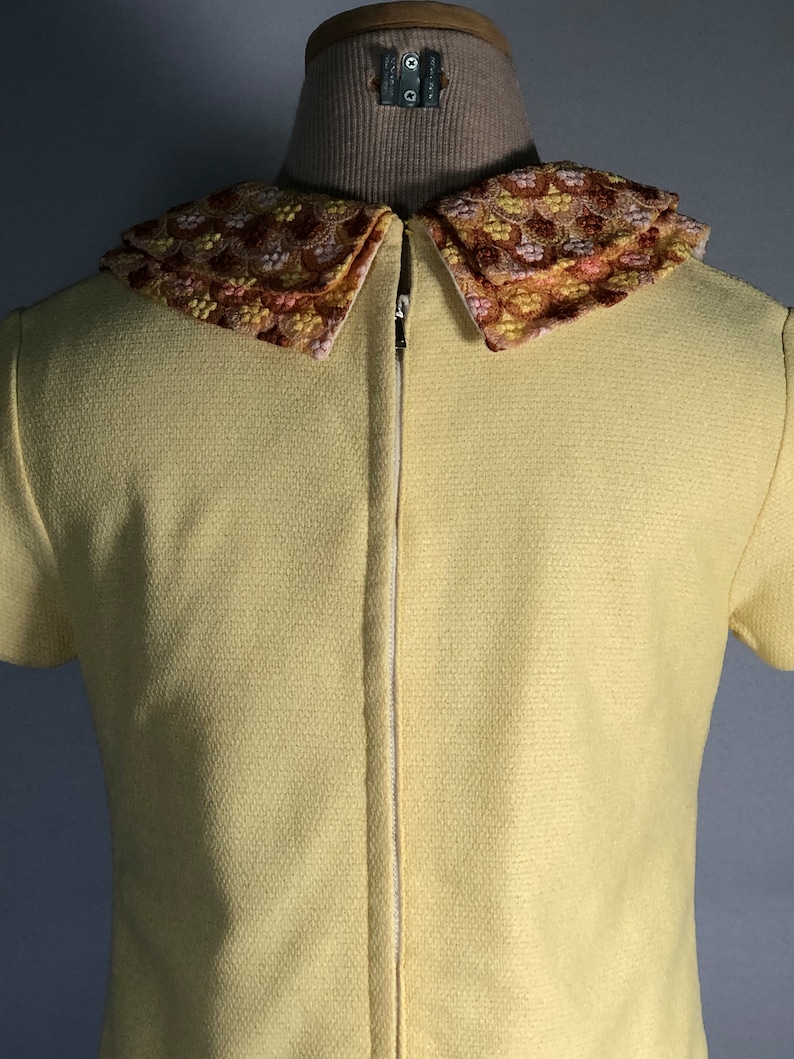 May include: A yellow short-sleeved dress with a large, embroidered collar. The collar is a multi-colored floral pattern in shades of pink, orange, and brown.