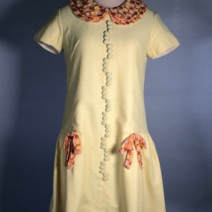May include: A yellow short-sleeved dress with a large, floral-patterned collar and matching bows on the pockets. The dress has a row of large, white buttons down the front.