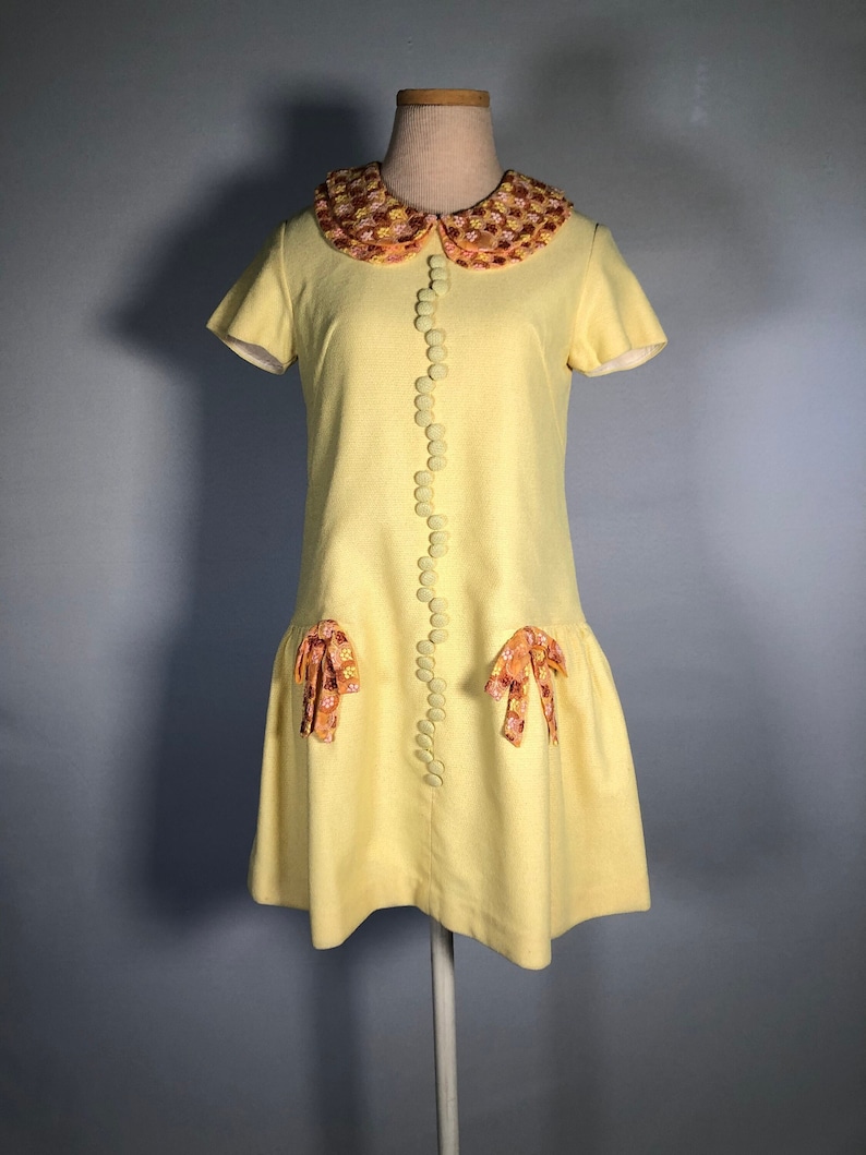May include: A yellow vintage dress with a floral print collar and matching bows on the skirt. The dress has a button-up front and short sleeves.