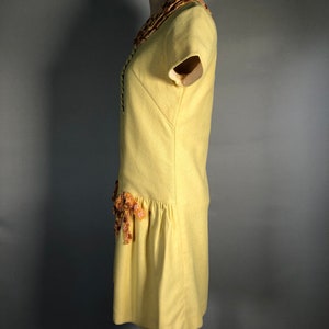 May include: A yellow short-sleeved dress with a floral trim around the neckline and a floral accent on the skirt.