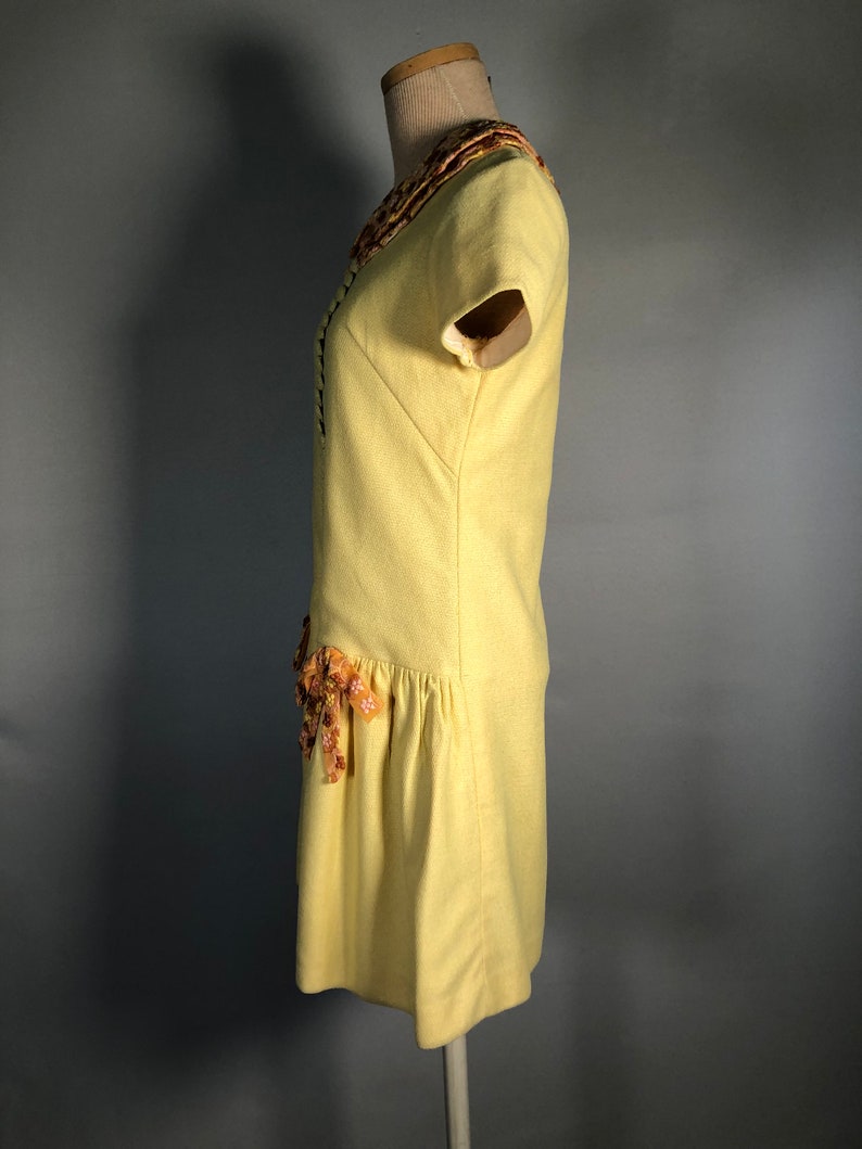 May include: A yellow short-sleeved dress with a floral trim around the neckline and a floral accent on the skirt.