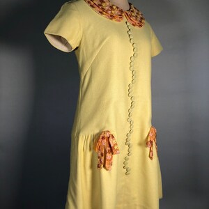 May include: A yellow vintage dress with a floral print collar and matching bows on the pockets. The dress has a button-up front and short sleeves.
