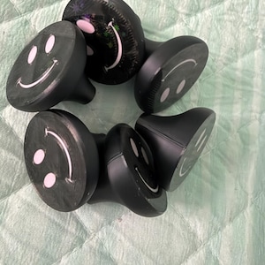 May include: A collection of five black drawer knobs, each featuring a circular face with a white printed smiley face design. The knobs have a smooth, matte finish and a tapered base, with one knob displaying a subtle glittery texture on its surface.