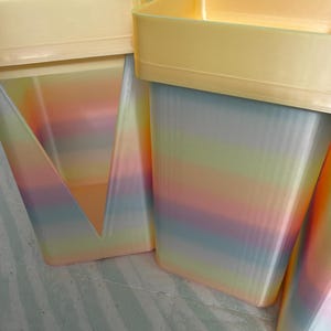 May include: A collection of rectangular containers featuring a pastel rainbow gradient pattern on the sides and a solid pale yellow rim at the top. The containers have a smooth finish and a structured, upright design with clean edges.