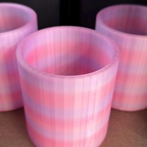 May include: Three cylindrical cups featuring a ribbed texture and a gradient color pattern that transitions between soft pink and light purple horizontal stripes. The cups have an open top and a smooth, rounded rim, displaying a consistent design.