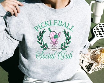 May include: A gray sweatshirt with a green and pink graphic of a cocktail glass with a lime and a cherry. The graphic is surrounded by a green wreath and the text "Pickleball Social Club".