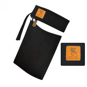 May include: Black fabric accessory with a rectangular shape and a strap. Features a small, square, tan-colored patch with a logo and Arabic text. Another similar patch is on the top part of the accessory. The accessory is likely used for fashion or personal care.