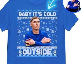 Może przedstawiać: Koszulka w kolorze królewskiego błękitu z białym wzorem śnieżynek i napisem "Baby it's cold outside". Na środku koszulki znajduje się twarz osoby.