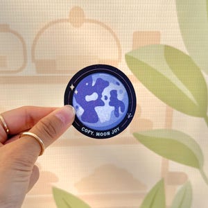 May include: A round sticker featuring a blue and white moon design with the text "COPY. MOON JOY" in white. The sticker is held by a hand with gold rings. The background is a light beige color with a leaf design.