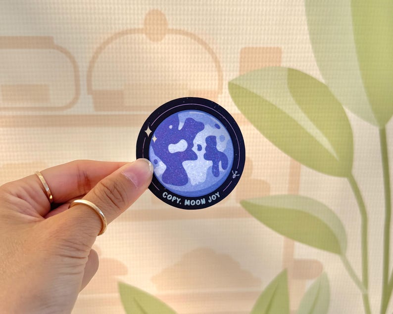 May include: A round sticker featuring a blue and white moon design with the text "COPY. MOON JOY" in white. The sticker is held by a hand with gold rings. The background is a light beige color with a leaf design.