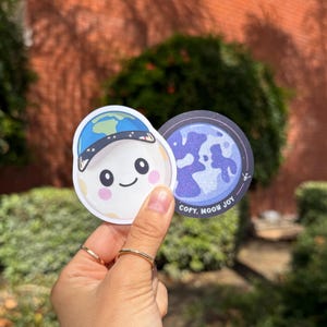 May include: Two stickers featuring a cartoon moon character wearing a blue hat with a world map design. The moon has rosy cheeks and a smiling face. The second sticker is a blue and purple graphic of the moon with the text "COPY. MOON JOY."