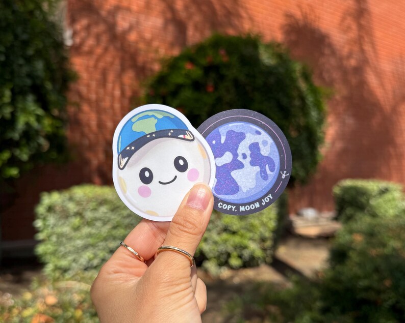 May include: Two stickers featuring a cartoon moon character wearing a blue hat with a world map design. The moon has rosy cheeks and a smiling face. The second sticker is a blue and purple graphic of the moon with the text "COPY. MOON JOY."