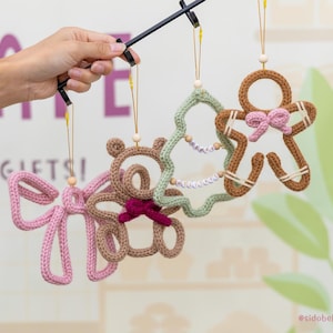 May include: Four crocheted ornaments hanging from a black display rack. The ornaments include a light pink bow, a tan bear with a dark pink bow, a light green tree with letter beads spelling HAPPY HOLIDAYS, and a brown gingerbread person.