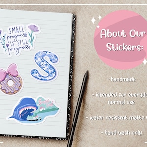 May include: A spiral-bound notebook with several colorful stickers. The stickers include a floral design with the text "Small progress is still progress", a blue letter "S", and a donut with a pink bow.