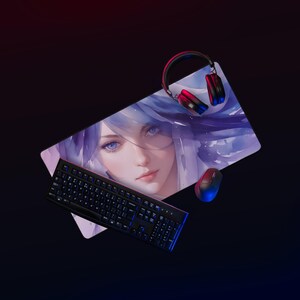 Op de afbeelding: Een gaming-opstelling met een grote rechthoekige muismat met een aquarel illustratie van een vrouw met blauw haar en ogen. Een zwart toetsenbord, een muis en een headset zijn ook aanwezig.