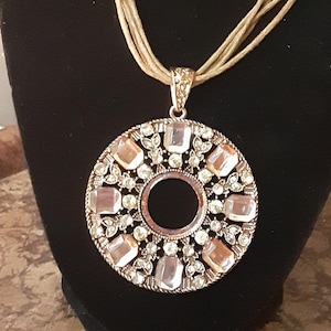 Könnte beinhalten: Eine mehrsträngige Halskette mit einem großen, runden Anhänger. Der Anhänger ist aus Metall und hat ein kreisförmiges Design mit klaren Strasssteinen.