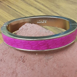 Op de afbeelding: Een gouden armband met een roze, getextureerd, krokodillenpatroon. De armband is gegraveerd met het woord "J.CREW".