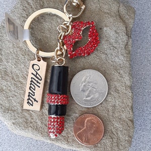 May include: Gold keychain with a red rhinestone lipstick charm, a red rhinestone lips charm, and a gold tag that says "Atlanta".